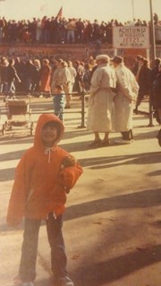 Image of young girl in red jacket (me) standing in front of Berlin Wall with people standing on top.
